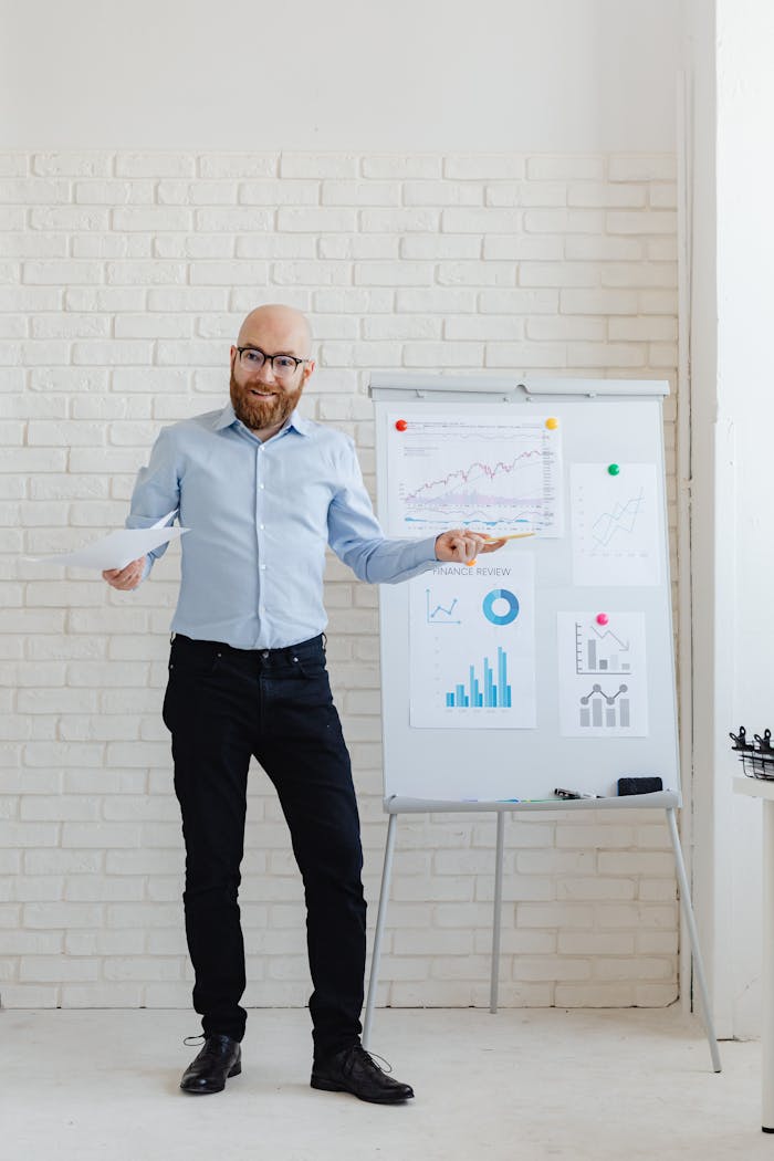 A bald, bearded man presents financial charts and graphs on a whiteboard.