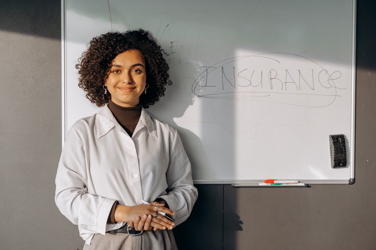 Smiling woman with curly hair standing by a whiteboard with insurance written on it, conveying professionalism.
