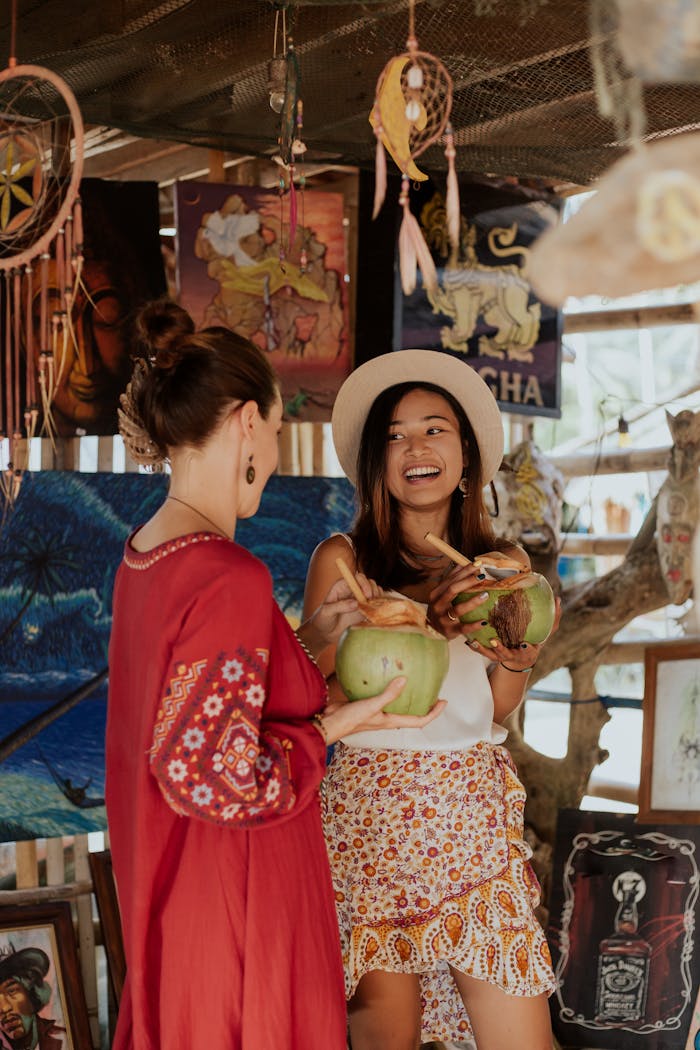 Two women smiling and enjoying coconut juice at an art market, surrounded by colorful paintings and decor.